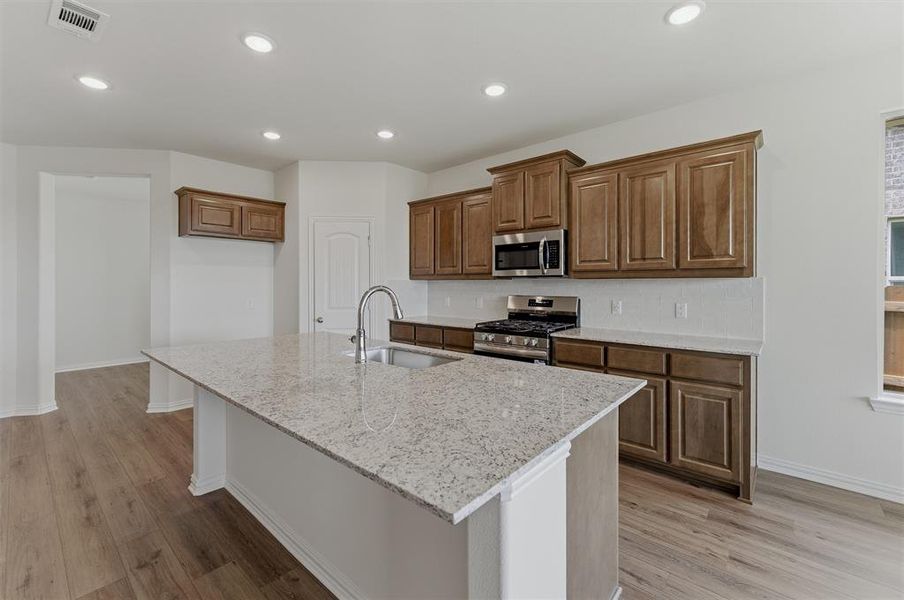 Kitchen featuring a large island with an undermount sink, granite-style countertop, stainless steel appliances, wood-finish cabinetry, and recessed lighting