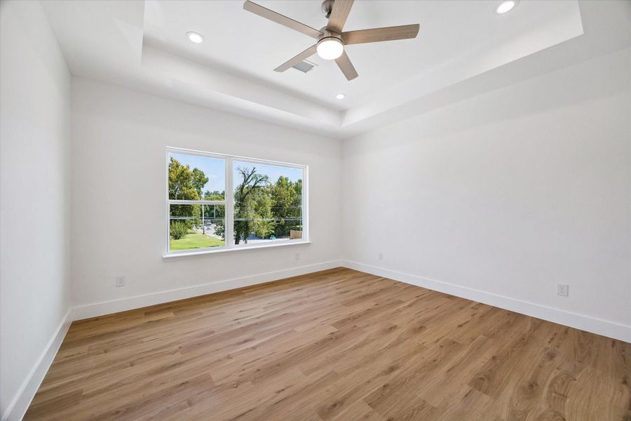 The spacious bedroom offers a bright and peaceful retreat with warm wood-look flooring, crisp neutral walls, and a large window overlooking mature greenery. A tray-style ceiling adds architectural interest, while recessed lighting and a contemporary ceiling fan enhance comfort. The generous dimensions allow room for a complete bedroom suite, additional seating, or a work-from-home area.