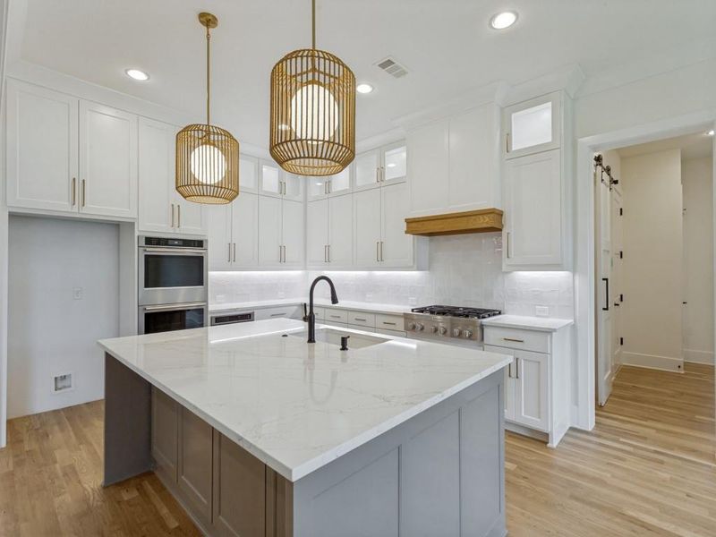 Modern kitchen featuring a large central island with a light-toned countertop, white cabinetry with gold-toned hardware, stainless steel appliances, and wood-finish flooring Modern kitchen featuring a large central island with a light-toned countertop, white cabinetry with gold-toned hardware, stainless steel appliances, and wood-finish flooring
