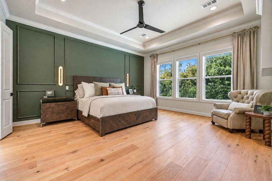 Spacious first-floor primary retreat features wood flooring, a dramatic tray ceiling and custom accent wall, with contemporary bedside lighting and a trio of oversized windows overlooking the private backyard.