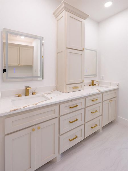 This bathroom features a double vanity with elegant marble countertops, gold fixtures, and ample cabinet storage. The bright, modern design is complemented by large mirrors and recessed lighting.