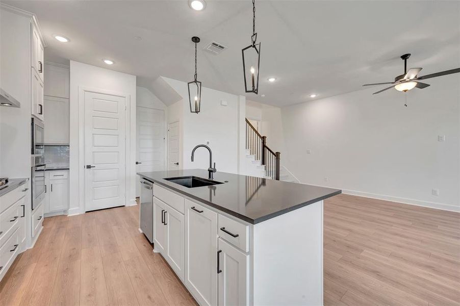 Open-concept kitchen featuring a large island with a dark countertop and sink, white cabinetry with black hardware, wood-finish flooring, and recessed lighting Open-concept kitchen featuring a large island with a dark countertop and sink, white cabinetry with black hardware, wood-finish flooring, and recessed lighting