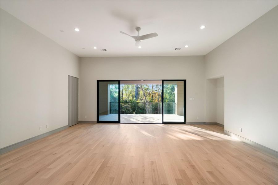 This spacious living area features light wood flooring, neutral walls, and a ceiling fan. Large sliding glass doors open to a balcony with a view of trees, providing ample natural light.