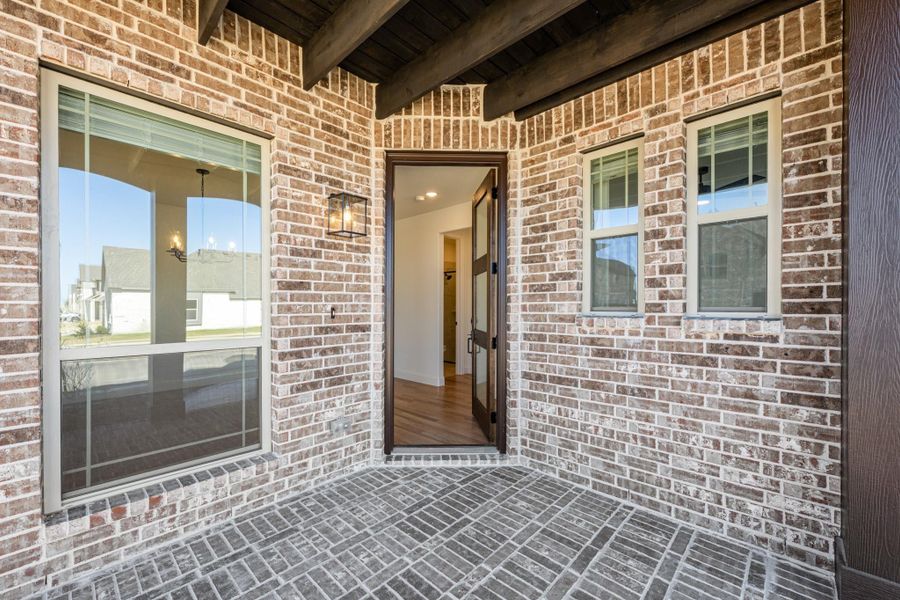 Covered brick porch with open front door, lantern light fixture, and exposed wood beam ceiling