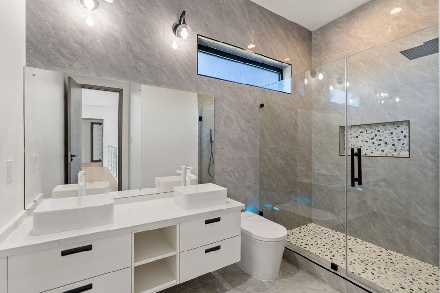This secondary bath features a double floating vanity with two vessel sinks while a glass-enclosed walk-in shower occupies the far end — gray tile with a mixed-stone floor tile in the shower pan that adds a layer of texture and contrast to the palette.