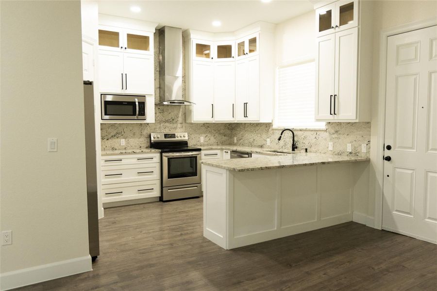 Kitchen with stainless steel appliances, white cabinetry, wall chimney range hood, light stone countertops, and a peninsula