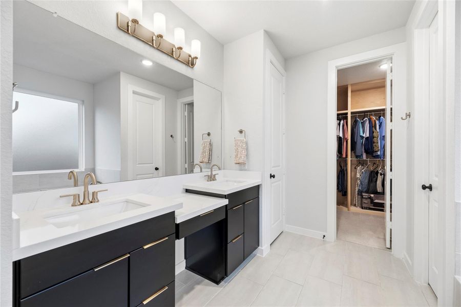 This modern bathroom features a double vanity with sleek cabinetry and gold fixtures, a large mirror with overhead lighting, and a spacious California walk-in closet. The space is bright and contemporary, with tile flooring and a frosted window for privacy.