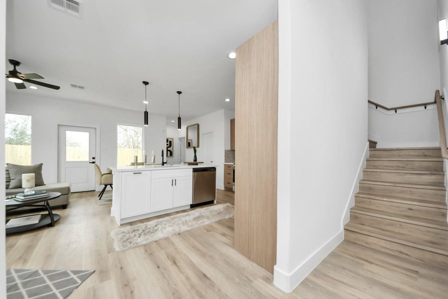 This photo showcases a modern open-concept living space with light wood flooring, a sleek kitchen island with pendant lighting, and a comfortable seating area. The room is brightened by natural light from large windows and a glass door leading outside. A staircase with wooden steps is visible, adding to the home's inviting and airy feel. This photo showcases a modern open-concept living space with light wood flooring, a sleek kitchen island with pendant lighting, and a comfortable seating area. The room is brightened by natural light from large windows and a glass door leading outside. A staircase with wooden steps is visible, adding to the home's inviting and airy feel.