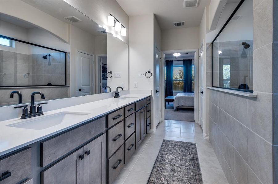 Ensuite bathroom featuring double vanity, healthy amount of natural light, light tile patterned floors, and tiled shower
