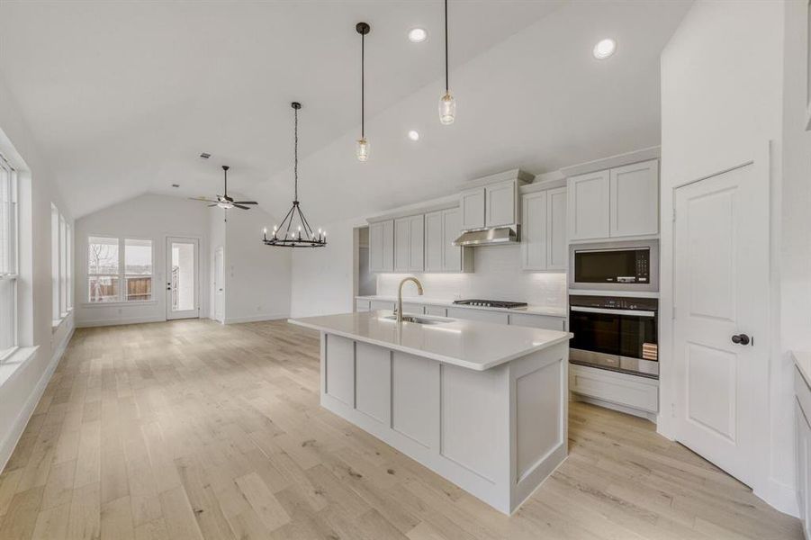 Kitchen featuring hanging lights, lofted ceiling, stainless steel appliances, an island with sink, and light wood-style floors Kitchen featuring hanging lights, lofted ceiling, stainless steel appliances, an island with sink, and light wood-style floors