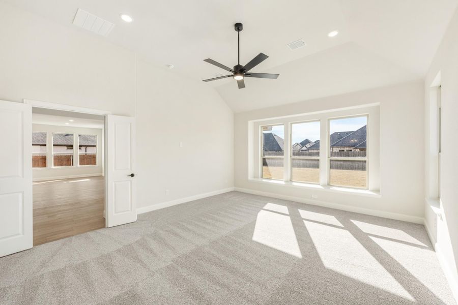 Empty bedroom with vaulted ceiling, ceiling fan, gray carpet, and triple windows overlooking neighborhood