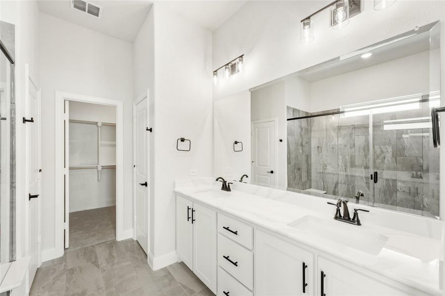 Bathroom with double vanity, white cabinets, large mirror, glass shower, and walk-in closet doorway visible