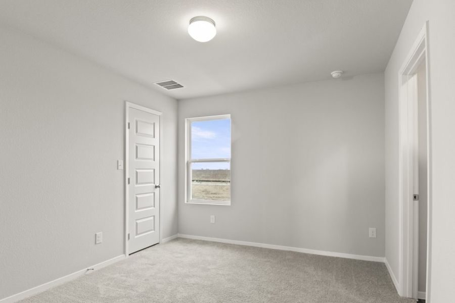 Image of a primary bedroom with tan carpeting and light grey walls with a window and a connecting closet and bathroom