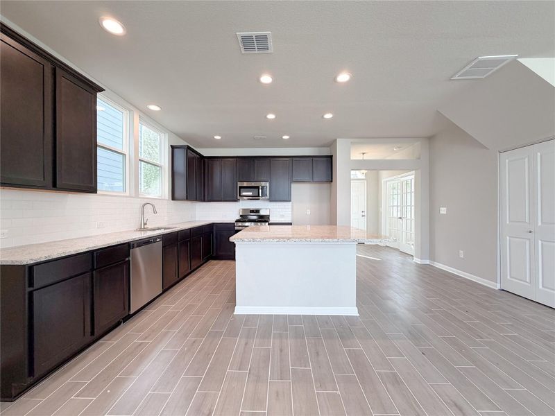 Open-concept kitchen featuring dark wood cabinetry, light-toned countertops, a white subway tile backsplash, and stainless steel appliances