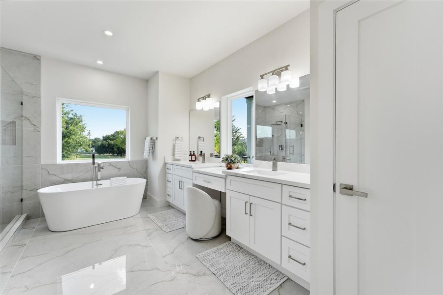 Spa-inspired primary bath featuring dual vanities, a dedicated makeup vanity, freestanding soaking tub, glass-enclosed shower, and elegant designer finishes. Spa-inspired primary bath featuring dual vanities, a dedicated makeup vanity, freestanding soaking tub, glass-enclosed shower, and elegant designer finishes.