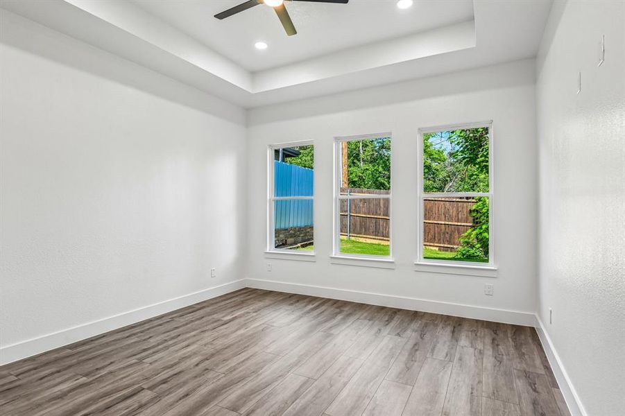 Room featuring wood-finish flooring, three large windows, a tray ceiling with recessed lighting, and a ceiling fan