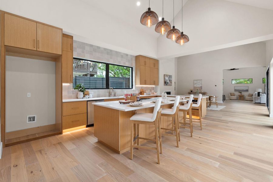 Open-concept kitchen featuring light wood-finish cabinetry, a large center island with seating, pendant lighting, high vaulted ceilings, and wood-finish flooring throughout