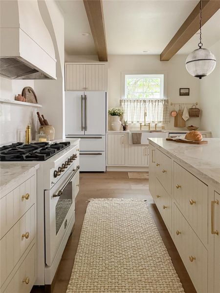 Gourmet kitchen featuring Forno professional-grade appliance package, custom made cabinetry with brass hardware, white oak flooring, a farmhouse sink, and exposed ceiling beams Gourmet kitchen featuring Forno professional-grade appliance package, custom made cabinetry with brass hardware, white oak flooring, a farmhouse sink, and exposed ceiling beams