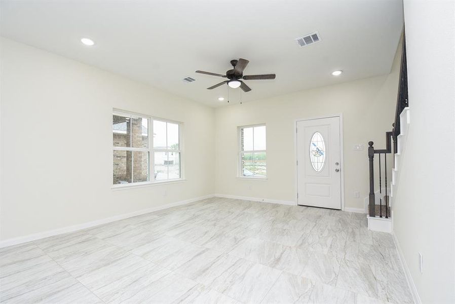 Large living room with tile flooring, a ceiling fan, windows, a front door with decorative glass, and the base of a staircase.