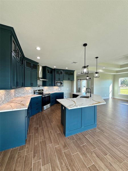 This kitchen features striking dark teal cabinetry, a large island with a marbled countertop, and modern stainless steel appliances. The open layout flows into a spacious living area with wood-look tile flooring, enhanced by ample natural light from large windows. This kitchen features striking dark teal cabinetry, a large island with a marbled countertop, and modern stainless steel appliances. The open layout flows into a spacious living area with wood-look tile flooring, enhanced by ample natural light from large windows.