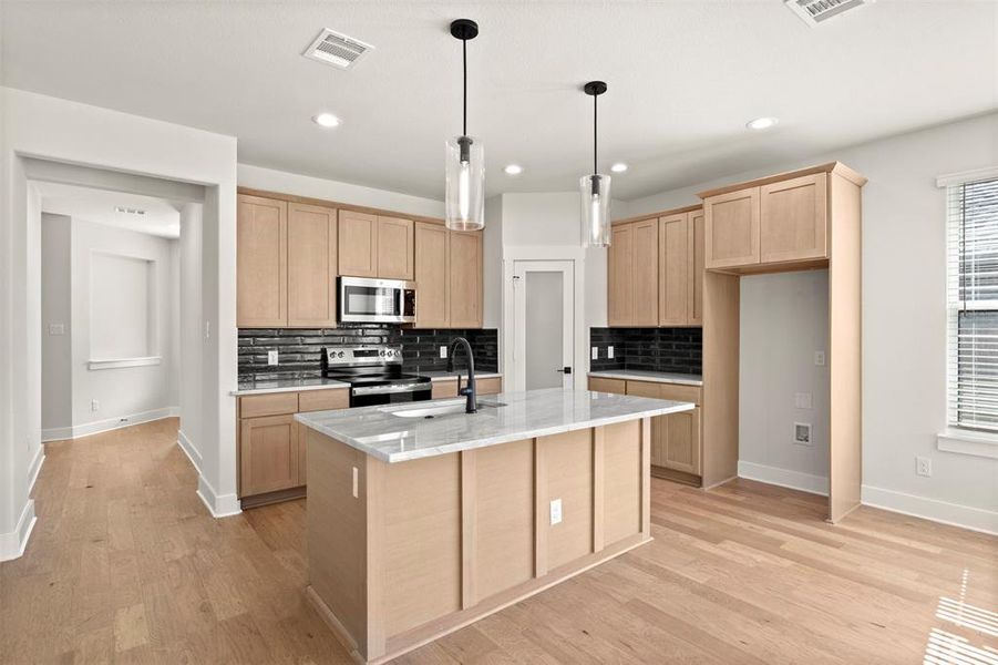 Kitchen with light brown cabinets, backsplash, light stone countertops, appliances with stainless steel finishes, and decorative light fixtures