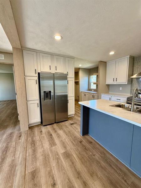 Kitchen featuring stainless steel refrigerator with ice dispenser, light countertops, light wood-style flooring, a textured ceiling, and white cabinetry Kitchen featuring stainless steel refrigerator with ice dispenser, light countertops, light wood-style flooring, a textured ceiling, and white cabinetry
