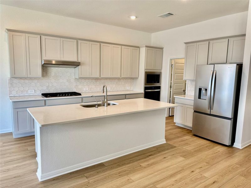 Kitchen featuring backsplash, black appliances, light stone countertops, light wood-type flooring, and a kitchen island with sink