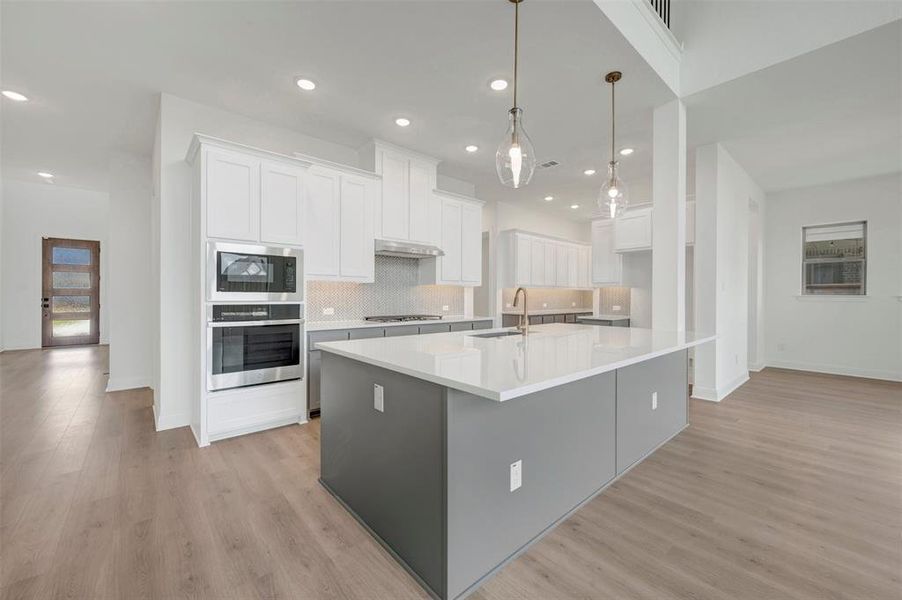 Contemporary kitchen featuring a large central island with a solid surface countertop, white shaker-style cabinetry, integrated stainless steel appliances, a tiled backsplash, and wood-finish flooring Contemporary kitchen featuring a large central island with a solid surface countertop, white shaker-style cabinetry, integrated stainless steel appliances, a tiled backsplash, and wood-finish flooring