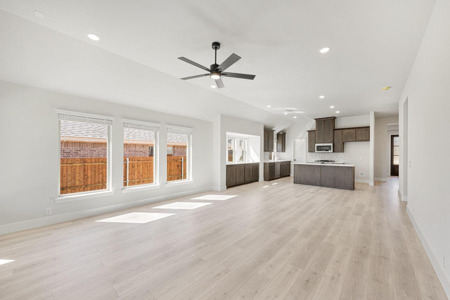 Open living area with light wood floors, ceiling fan, and kitchen with dark cabinets and island in background