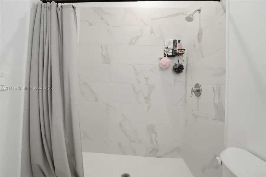 Shower enclosure featuring large-format light-toned stone-finish wall tile with prominent veining, a wall-mounted showerhead, and a single-handle pressure balance valve