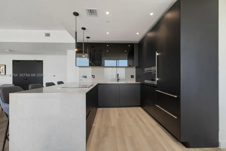Contemporary kitchen featuring custom dark cabinetry, stone-surfaced waterfall island, integrated built-in appliances, recessed lighting, and wood-finish flooring