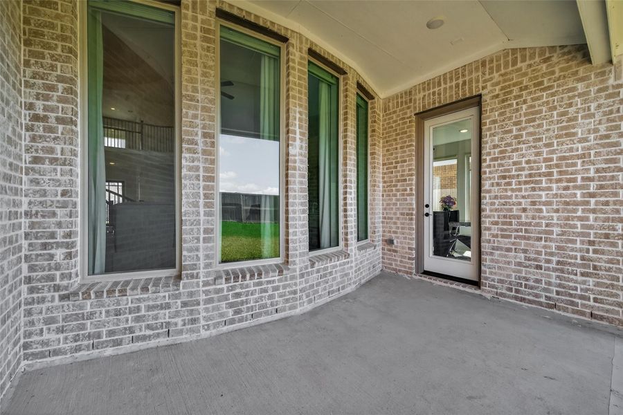 This photo shows a covered patio area with light brick walls and large, tall windows providing natural light. There's a glass door leading inside, and the space offers a clean, open area ideal for relaxation or entertaining.