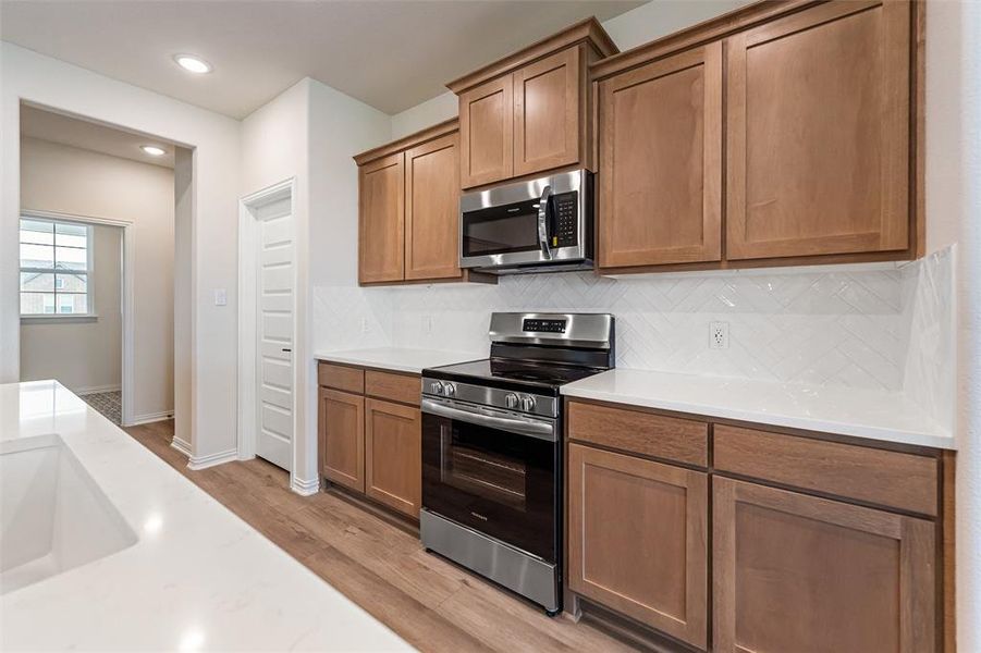 Kitchen with stainless steel appliances, brown cabinetry, light wood finished floors, tasteful backsplash, and light stone counters