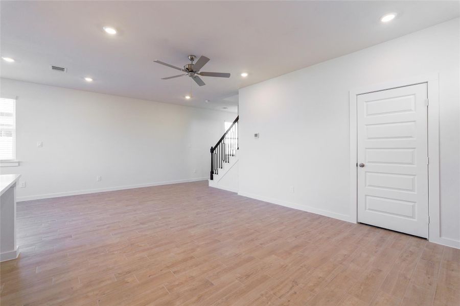 This photo shows a spacious, open-concept room with light wood flooring and neutral walls. It features recessed lighting, a ceiling fan, and a staircase with dark railings. A white door is present, enhancing the modern, clean design. This photo shows a spacious, open-concept room with light wood flooring and neutral walls. It features recessed lighting, a ceiling fan, and a staircase with dark railings. A white door is present, enhancing the modern, clean design.