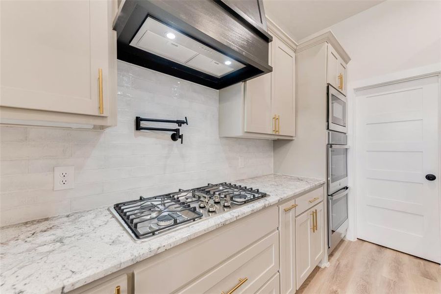 Kitchen with range hood, decorative backsplash, light stone counters, stainless steel appliances, and light wood-style floors Kitchen with range hood, decorative backsplash, light stone counters, stainless steel appliances, and light wood-style floors