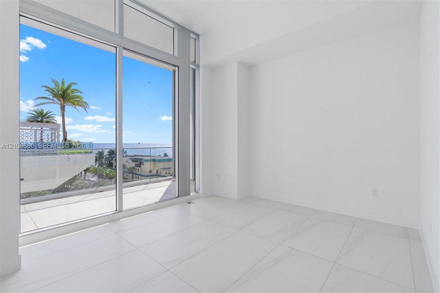 Floor-to-ceiling glass wall, large-format tile flooring, and sliding glass door opening to a private balcony with glass railing and water backdrop