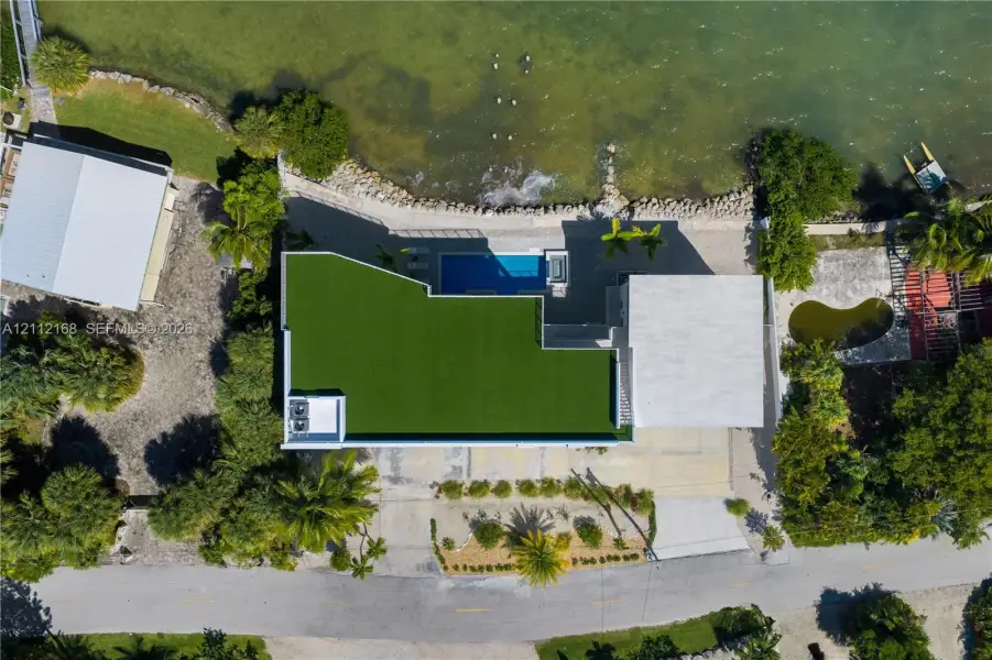Aerial perspective of waterfront property featuring a swimming pool, synthetic turf lawn, stone-reinforced shoreline, and paved driveway Aerial perspective of waterfront property featuring a swimming pool, synthetic turf lawn, stone-reinforced shoreline, and paved driveway