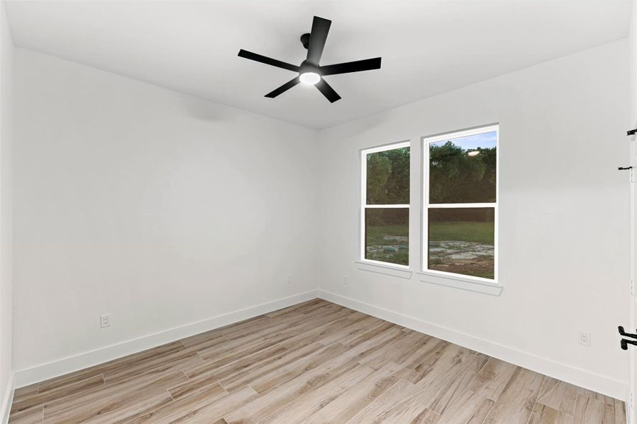 This room features a modern ceiling fan, light wood-like tile flooring, and large windows that provide ample natural light with views of greenery. The white walls create a bright, clean space. This room features a modern ceiling fan, light wood-like tile flooring, and large windows that provide ample natural light with views of greenery. The white walls create a bright, clean space.