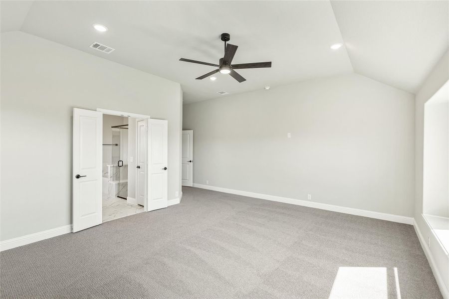 Spacious bedroom with vaulted ceiling, ceiling fan, gray carpet, and open doors leading to ensuite bathroom Spacious bedroom with vaulted ceiling, ceiling fan, gray carpet, and open doors leading to ensuite bathroom