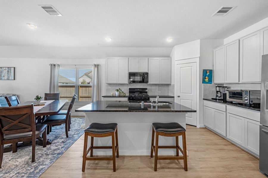 This light and bright kitchen features a large granite island, custom stained cabinets, a large sink, recessed lighting, and beautiful backsplash.
