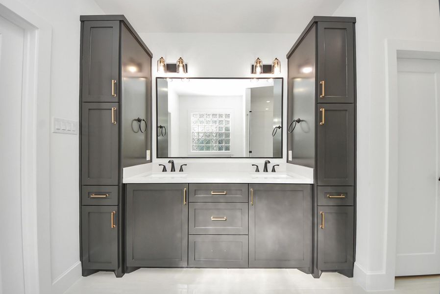 A sleek bathroom design highlighted by a modern double vanity with dark cabinetry and gold hardware. A large mirror and overhead lighting illuminate the space, balanced by clean white walls and countertops.