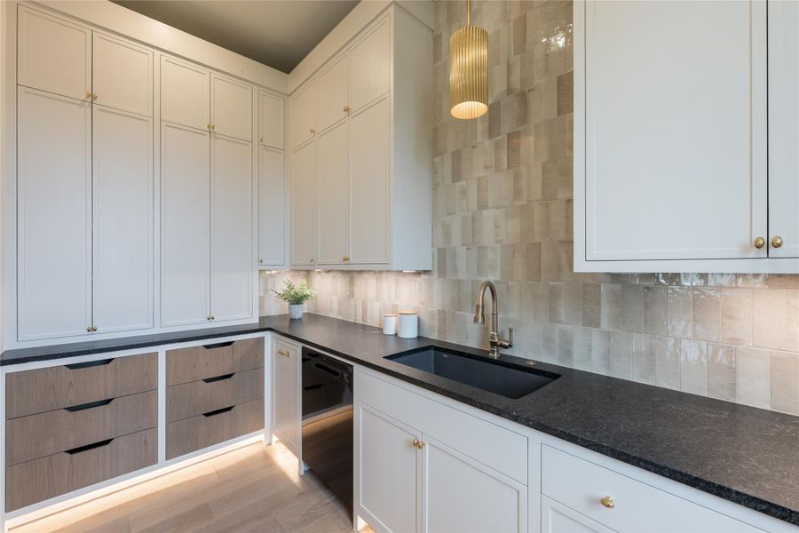 Another view pairs textural handmade tile with sleek black stone countertops and creamy cabinetry, all softly illuminated by under-cabinet lighting and a fluted pendant overhead. Custom drawer fronts in a contrasting wood tone add depth and craftsmanship, while the brass faucet and hardware echo the elegance found throughout the main kitchen. Another view pairs textural handmade tile with sleek black stone countertops and creamy cabinetry, all softly illuminated by under-cabinet lighting and a fluted pendant overhead. Custom drawer fronts in a contrasting wood tone add depth and craftsmanship, while the brass faucet and hardware echo the elegance found throughout the main kitchen.