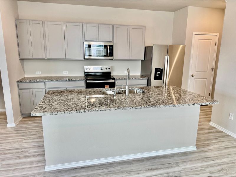 Kitchen with gray cabinets, stainless steel appliances, light stone counters, light wood-style floors, and a kitchen island with sink
