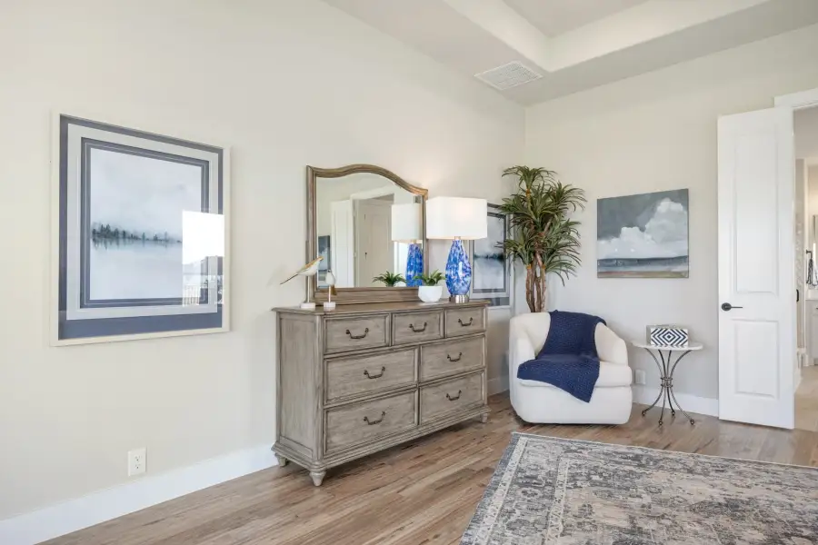 Bedroom sitting area with wood dresser, mirror, white accent chair, and hardwood floors under a tray ceiling Bedroom sitting area with wood dresser, mirror, white accent chair, and hardwood floors under a tray ceiling