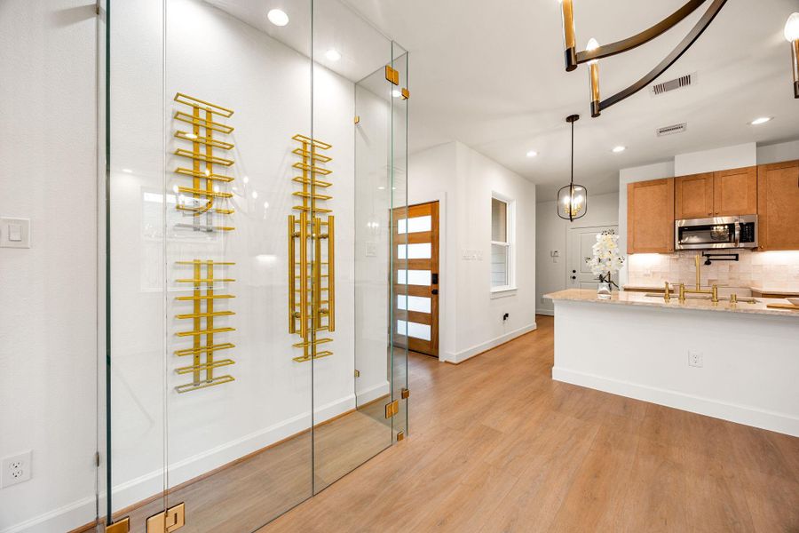 A striking glass-enclosed wine room with sleek metal racking creates a bold architectural statement, seamlessly integrated into the main living area. Warm wood flooring, designer lighting, and clean modern lines flow effortlessly into the adjacent kitchen, offering a sophisticated space ideal for entertaining and everyday living.