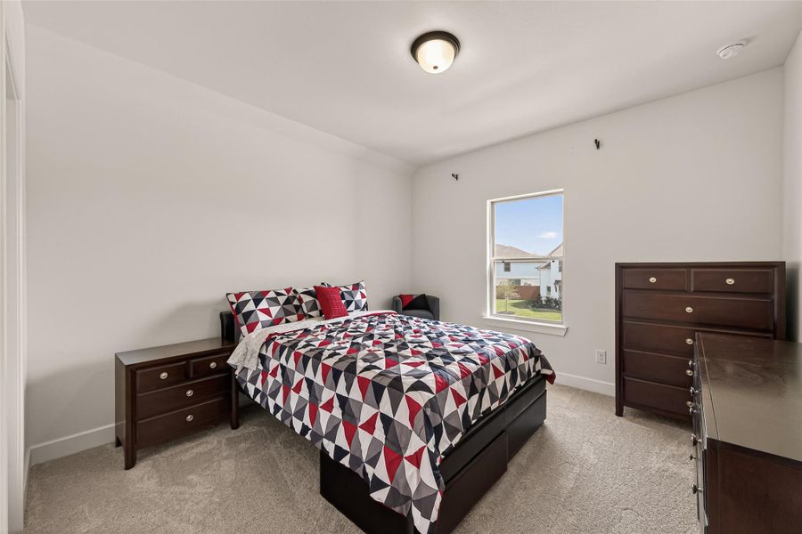 This bedroom features neutral walls, plush carpeting, and a window offering natural light. It includes a bed with a modern geometric bedspread, a nightstand, and a dresser, creating a functional and inviting space.