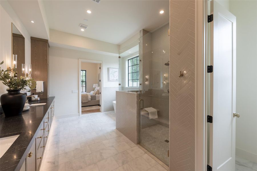 The view from this angle captures the intentional flow between the primary suite and spa-inspired bath. Anchored by a dual-sink vanity with honed black stone counters, brass fixtures, and oak cabinetry, the space is both elevated and inviting. The walk-in shower features a built-in bench, frameless glass, and a continuous herringbone tile that wraps the walls with soft texture. The view from this angle captures the intentional flow between the primary suite and spa-inspired bath. Anchored by a dual-sink vanity with honed black stone counters, brass fixtures, and oak cabinetry, the space is both elevated and inviting. The walk-in shower features a built-in bench, frameless glass, and a continuous herringbone tile that wraps the walls with soft texture.