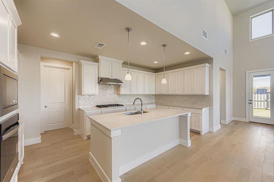 Kitchen featuring backsplash, appliances with stainless steel finishes, light wood-type flooring, decorative light fixtures, and a high ceiling