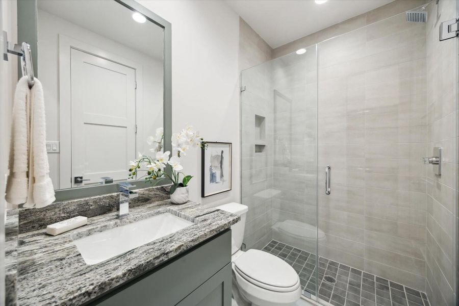 This modern bathroom features a sleek design with a glass-enclosed shower, marble countertop, and neutral tilework. It includes a large mirror, stylish fixtures, and a touch of elegance with a floral arrangement.