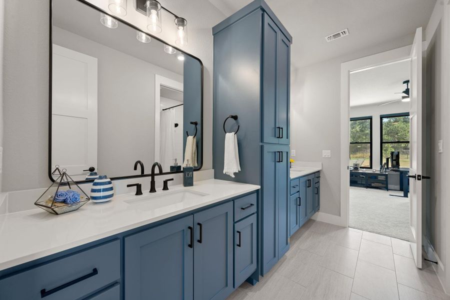A spacious bathroom features dual vanities and custom blue cabinetry alongside sleek modern tile flooring.